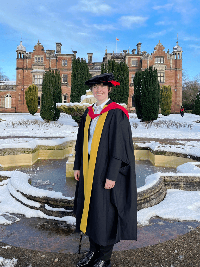 me on my graduation day in a full PhD gown and cap in front of an old building